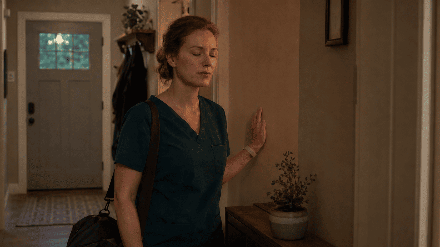 A nurse in scrubs stands at a kitchen counter looking at her phone after a long ICU shift, while a child sits blurred in the background — depicting the post-shift irritability and emotional distance that can affect nurse families.