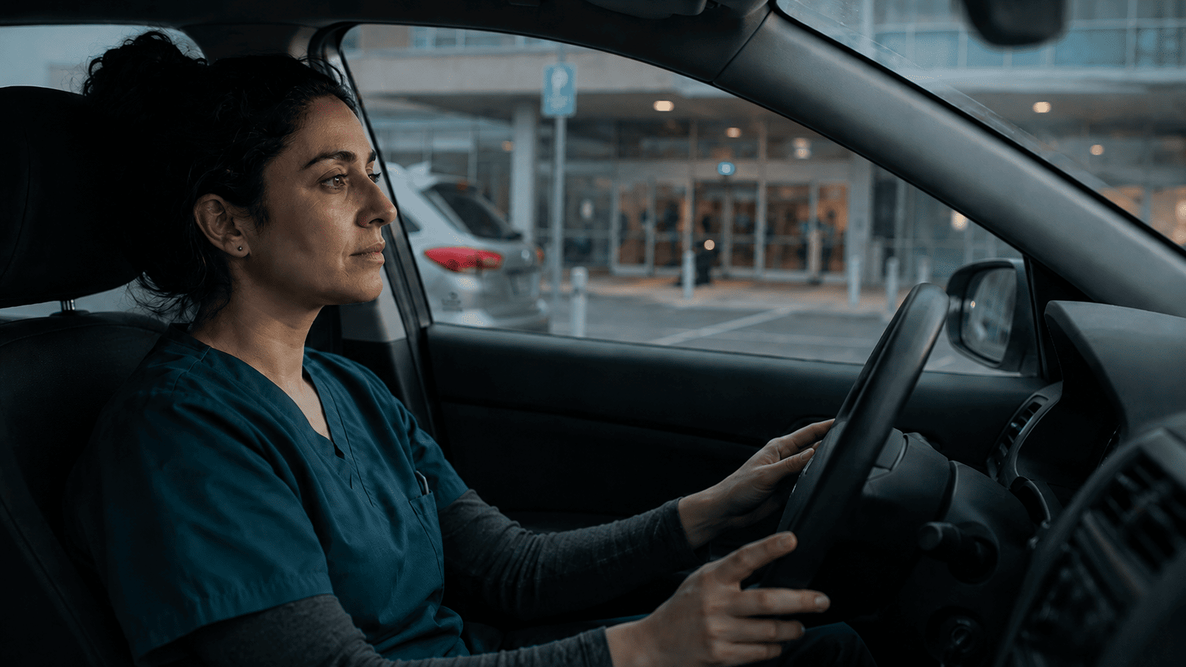 A nurse in scrubs sits in a parked car with both hands on the steering wheel, looking toward a hospital entrance with a still, bracing expression, experiencing pre-shift dread before an ICU or NICU shift.